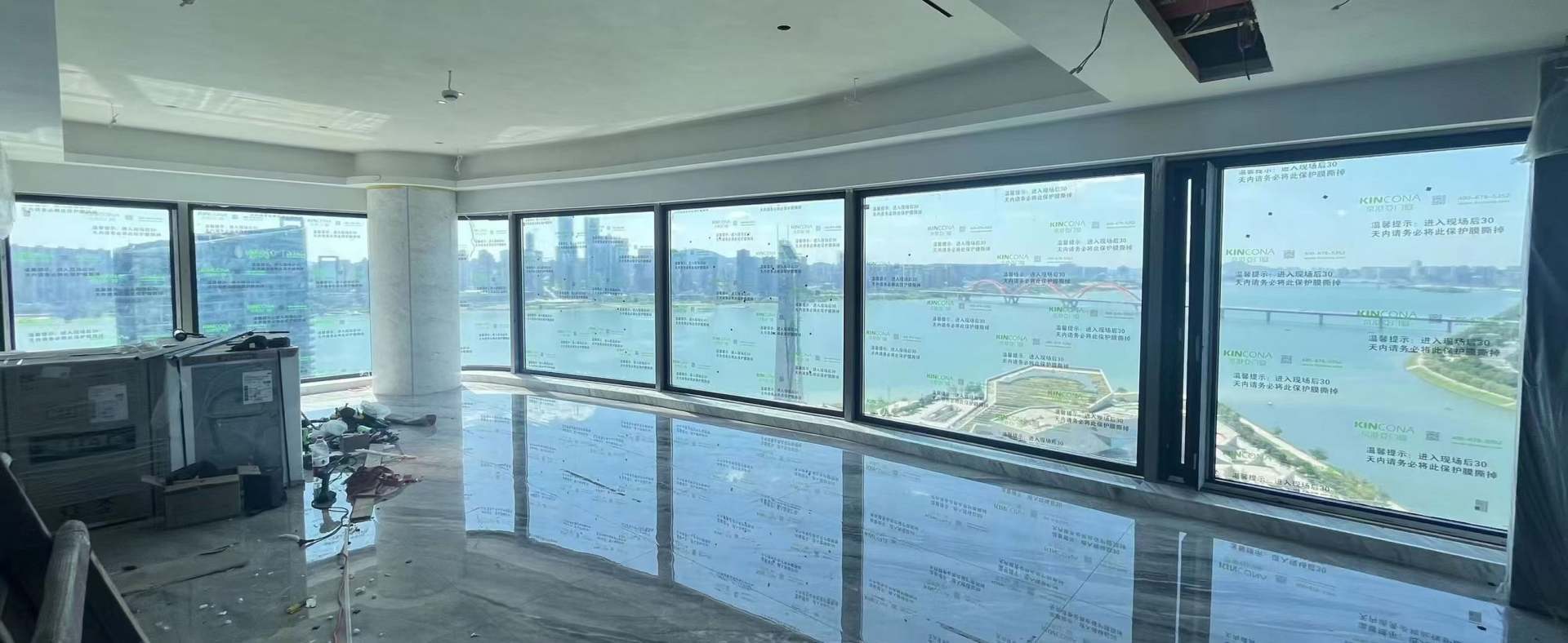 这是一张高层江景房的室内施工现场实拍图。画面核心是一排宽大的落地窗，窗外可见宽阔江面与跨江大桥，视野极佳。窗户玻璃上贴有“KINCONA 京港亚门窗”的品牌保护