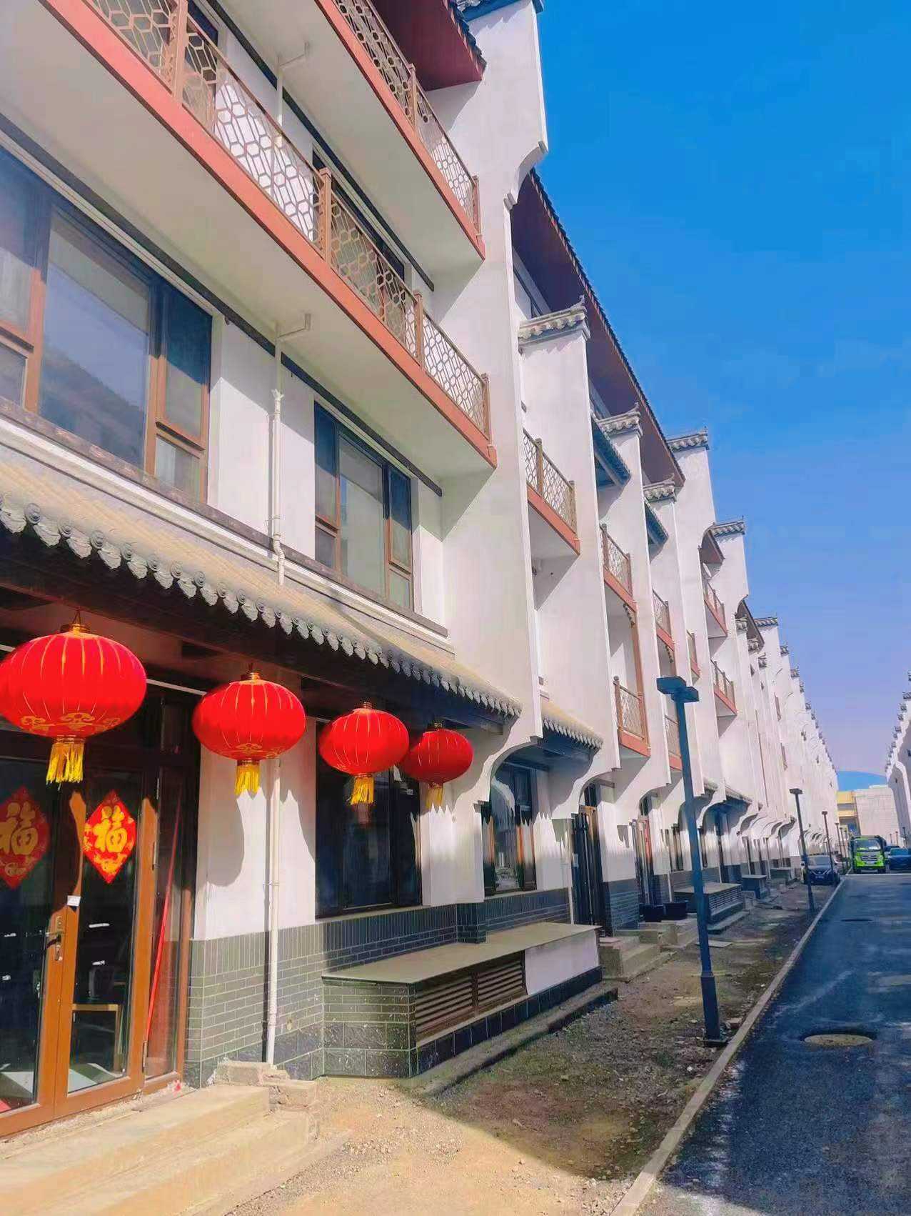 户外实景拍摄的一排新中式联排别墅，蓝天映衬下白墙黛瓦格外醒目。建筑融合传统飞檐翘角与现代大玻璃窗设计，阳台配有红棕色几何纹样金属栏杆。底层采用深青色砖块墙裙，屋