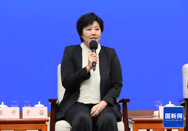 青い背景の会場でマイクを持ち、椅子に座る春節晩会総監督の于蕾。